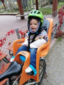 1-year old toddler on a baby seat attached to a bike. Kid's awkwardly dressed in warm clothes, and very cute.